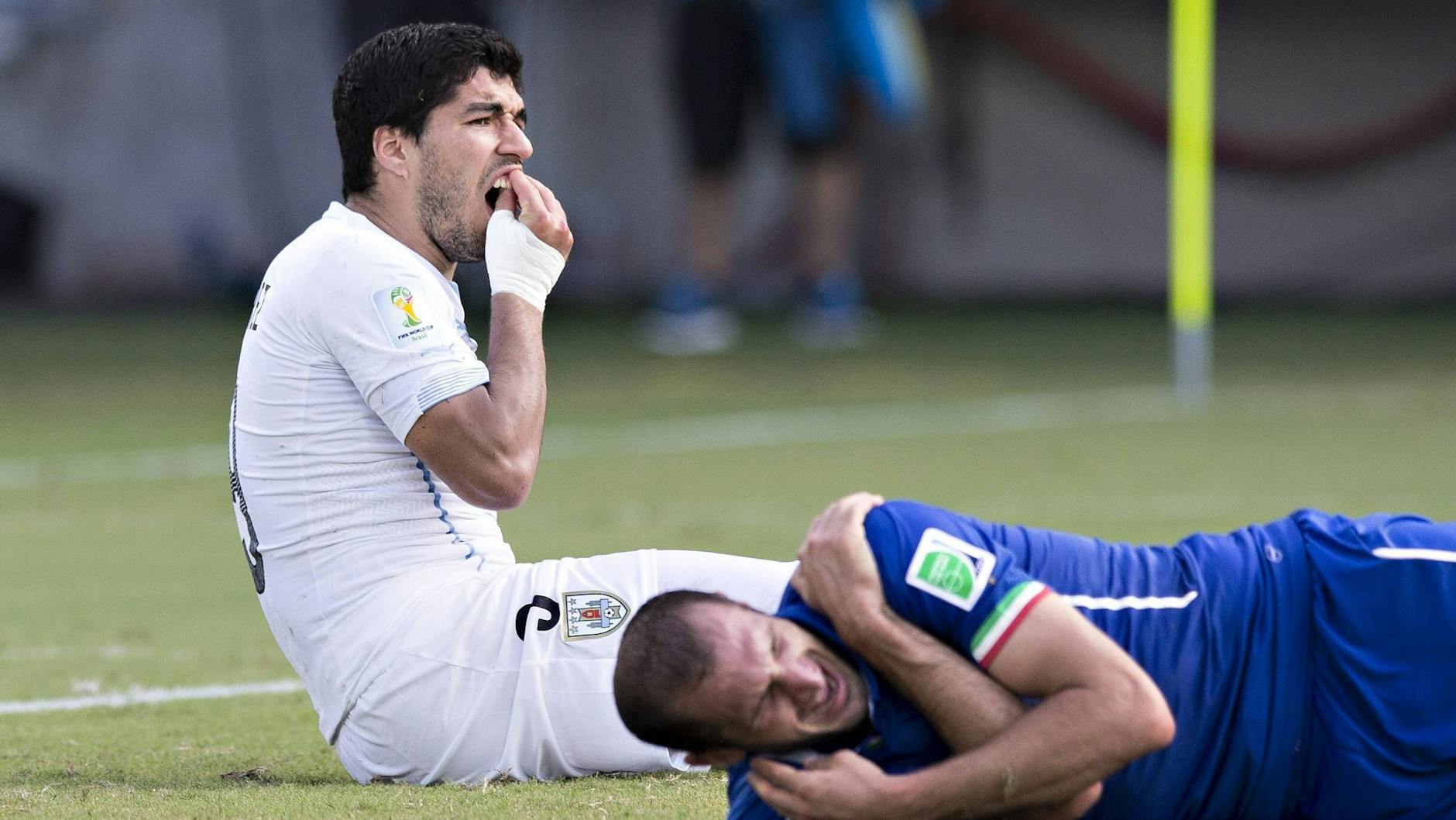 Uruguays Luis Suárez kontrolliert nach seiner Beißattacke bei der WM 2014 die Zähne, Italien-Star Giorgio Chiellini krümmt sich vor Schulter-Schmerzen auf dem Rasen.